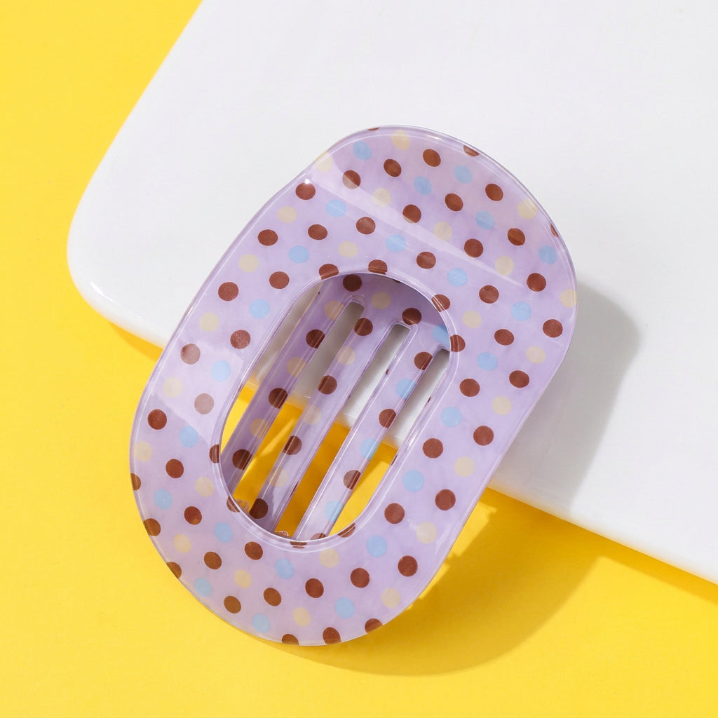 A translucent lavender hair claw clip with brown, light blue, and yellow polka dots, displayed on a bright yellow background with a white geometric prop.