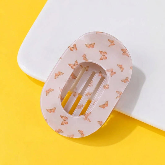 An oval-shaped translucent hair claw clip with a delicate butterfly pattern, resting on a white rectangular block against a bright yellow background. 
