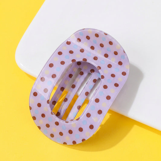 A translucent lavender hair claw clip with brown, light blue, and yellow polka dots, displayed on a bright yellow background with a white geometric prop. 