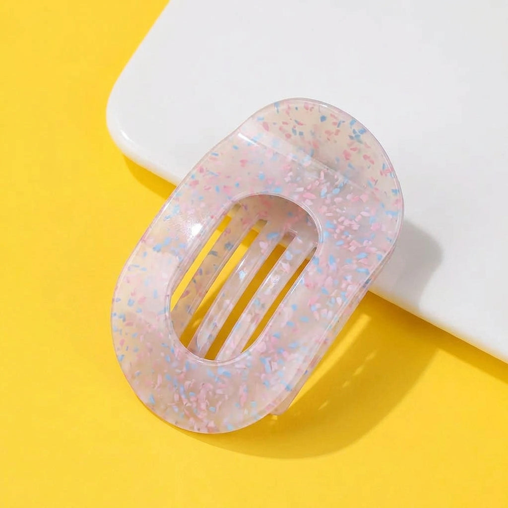 A translucent oval hair claw clip with a pink and light blue floral pattern, displayed on a bright yellow background with a white geometric prop. 